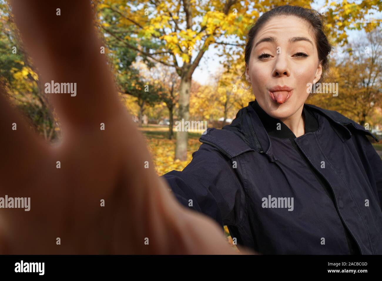 Freche junge Frau in ihrem 20s die Zunge heraus klemmt unter selfie Foto im Park mit Herbstfarben posing - Wide Angle Shot Holding unsichtbare Smartphone mit ausgestrecktem Arm Stockfoto