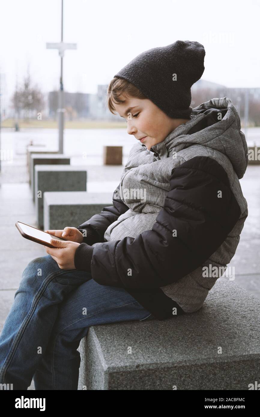 7 Jahre alten Jungen mit Smartphone Handy draußen sitzen allein auf der Straße im Winter, Entsättigung der Farben Stockfoto