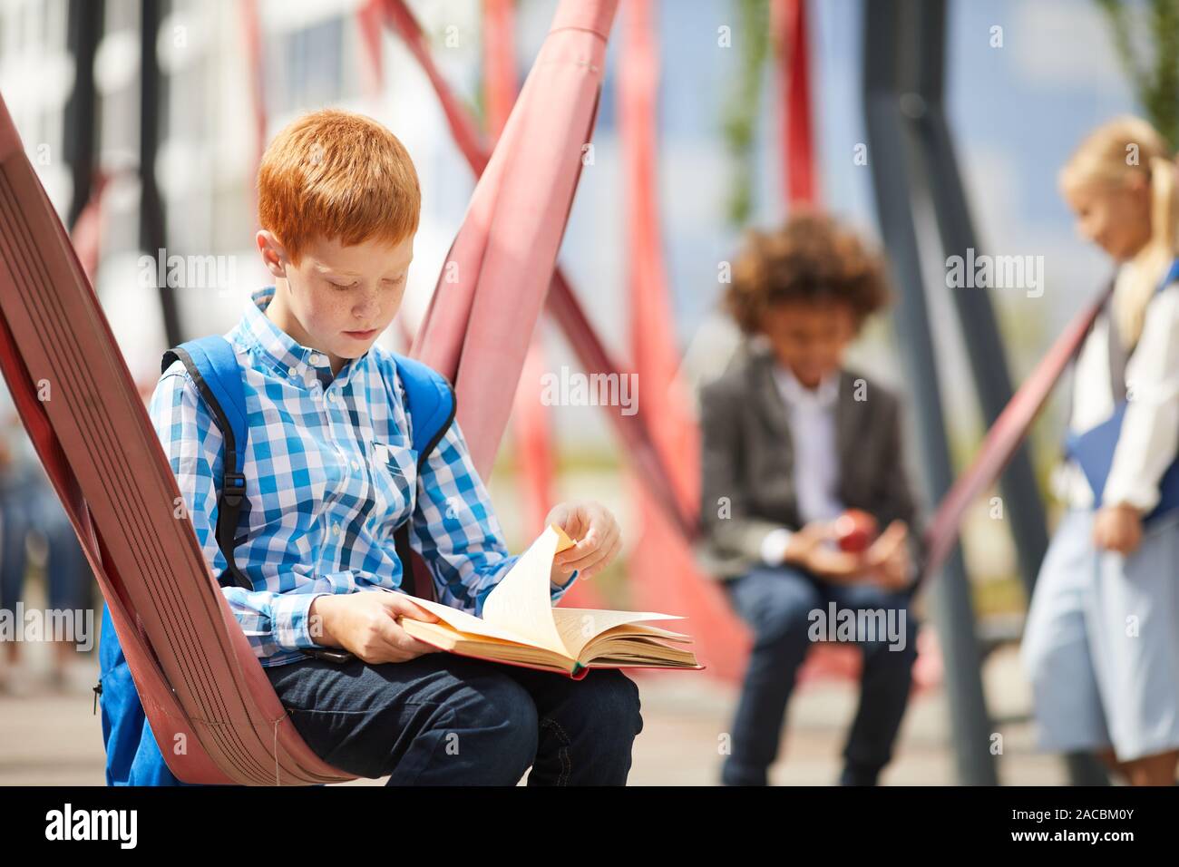Schüler mit Rucksack auf dem Rücken sitzen auf der Schaukel und ein Buch auf dem Schulhof Stockfoto
