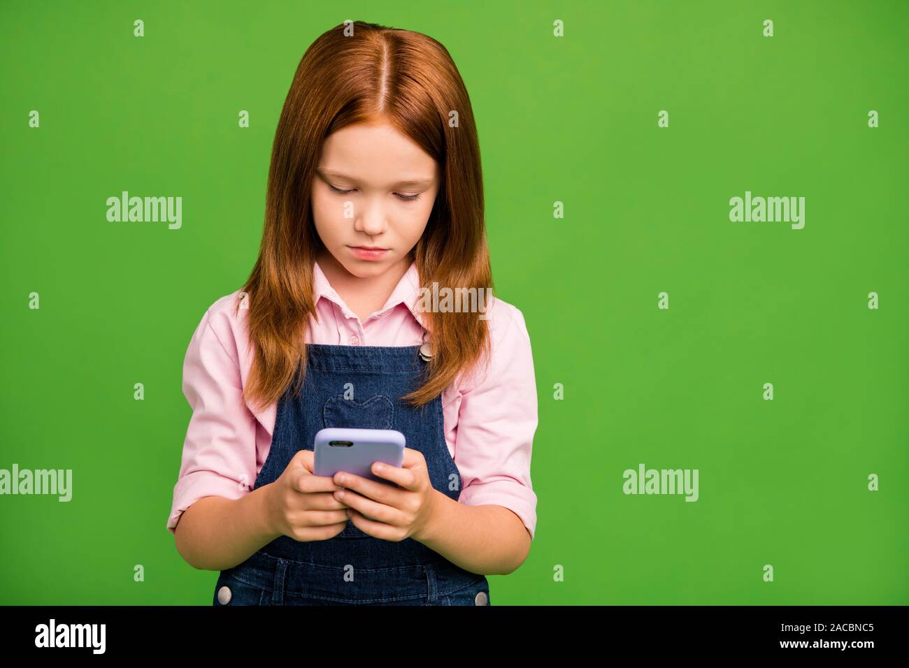 Foto des hübschen kleinen Schulkind holding Telefon Hände suchen Hausaufgaben zusätzliche Informationen tragen Jeans overall rosa Shirt isoliert Grün Stockfoto