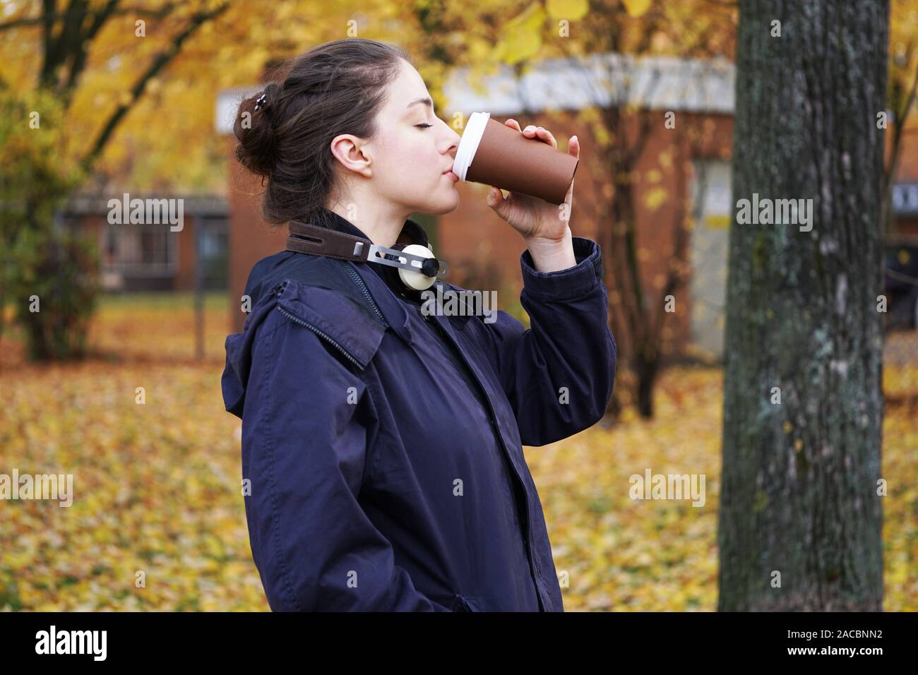 Trendige junge Frau hipster tragen parka Mantel mit Kopfhörer um den Hals und das Trinken von Kaffee Tasse zu beim Gehen im Park an einem kalten Herbsttag. Stockfoto
