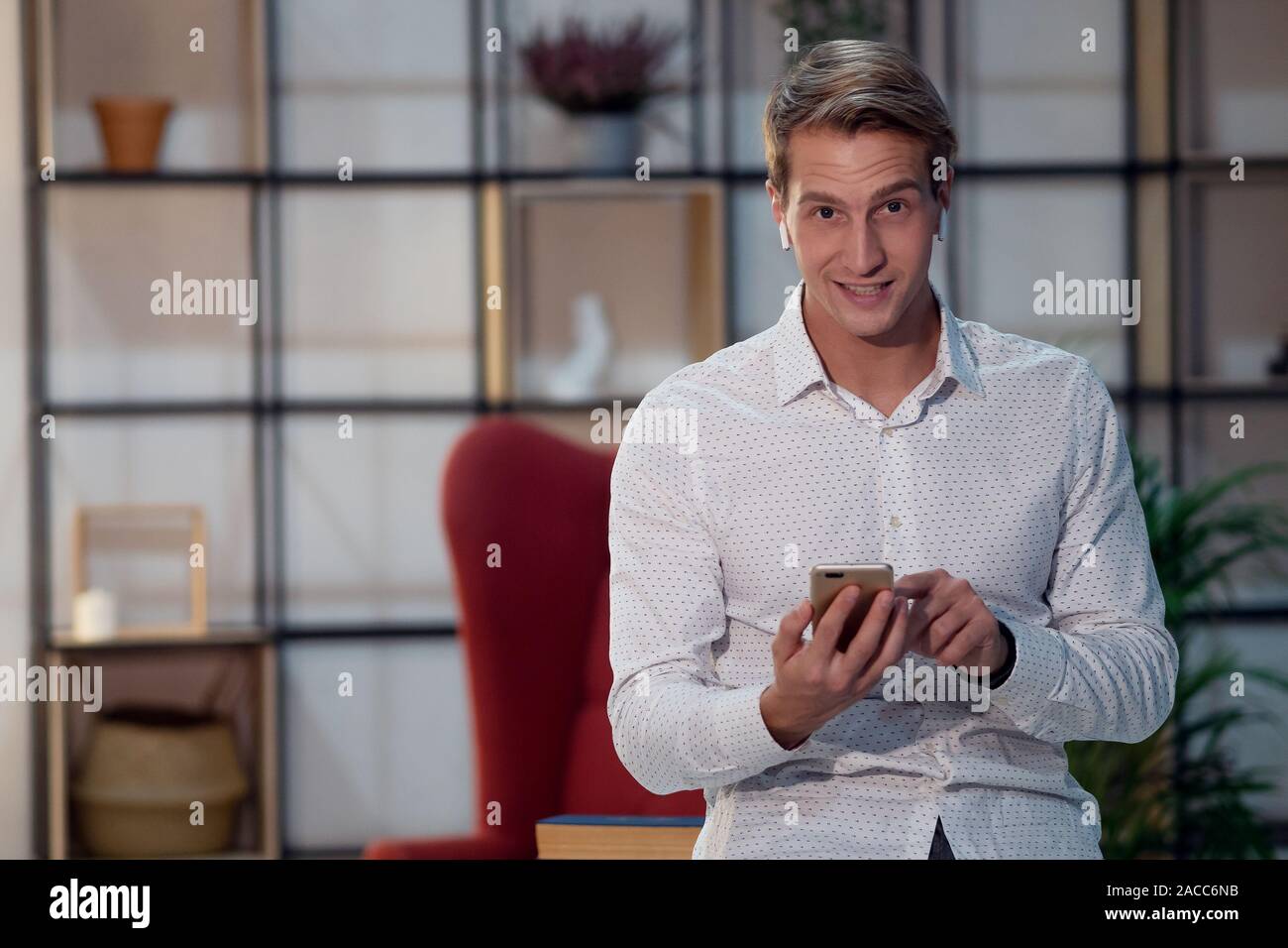 Lächelnder junger Mann in kabellosen Kopfhörern, Portrait im Loft-Interieur, stilvolles Zimmer. Positiver Kerl verwendet Smartphone für Online-Schulungen. Stockfoto