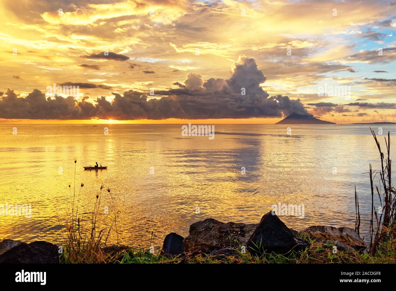 Farbige Himmel bei Sonnenuntergang mit Person in einem Boot, markanten Wolkenbildung und Berg im Hintergrund, Pflanzen und Steine im Vordergrund - Ort: Stockfoto