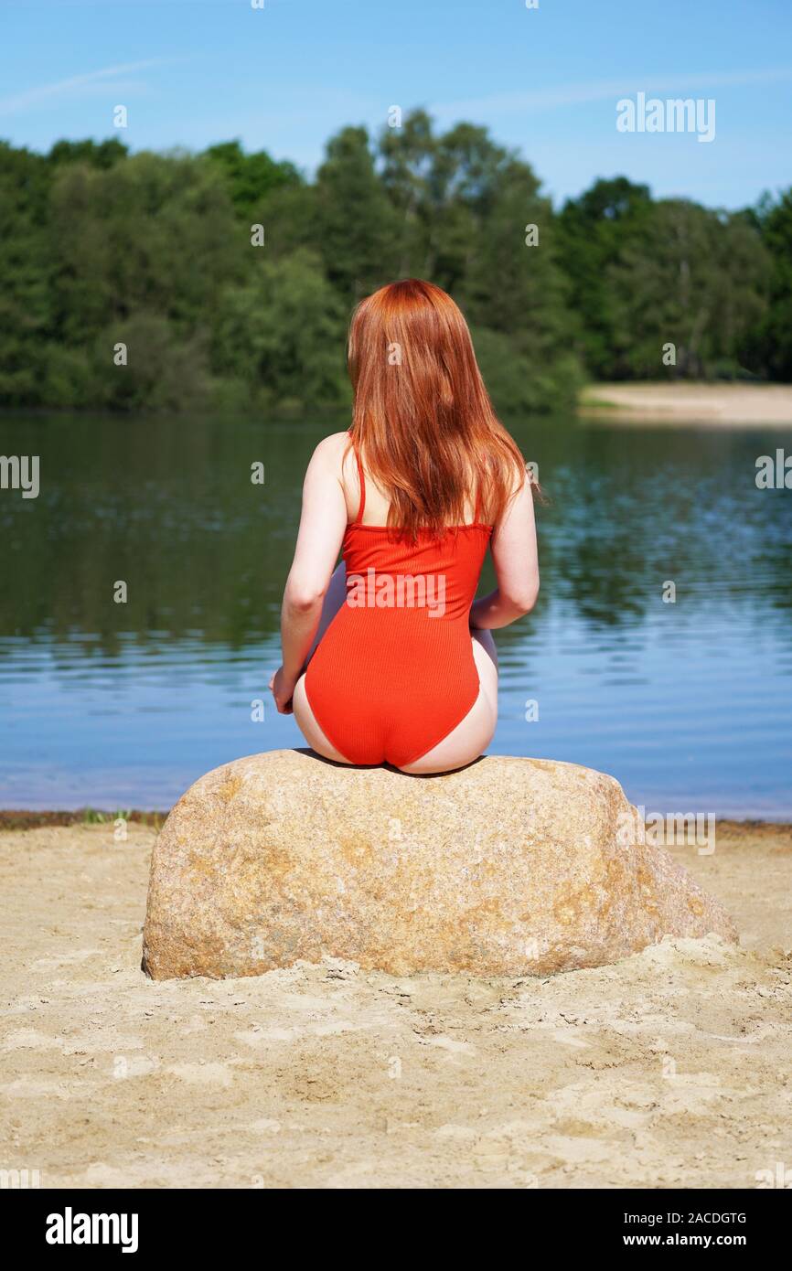 Ansicht der Rückseite des jungen Frau rote Haare und Badeanzug oder Badehose sitzen auf Rock am Sandstrand am idyllischen See - Sommer lifestyle Stockfoto