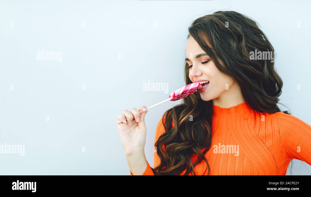 Junge schöne Mädchen mit dunklen lockigen Haar isst eine rosa Lollipop candy schreien Stolz in einem hellen orange Pullover auf einem einfachen Hintergrund Stockfoto