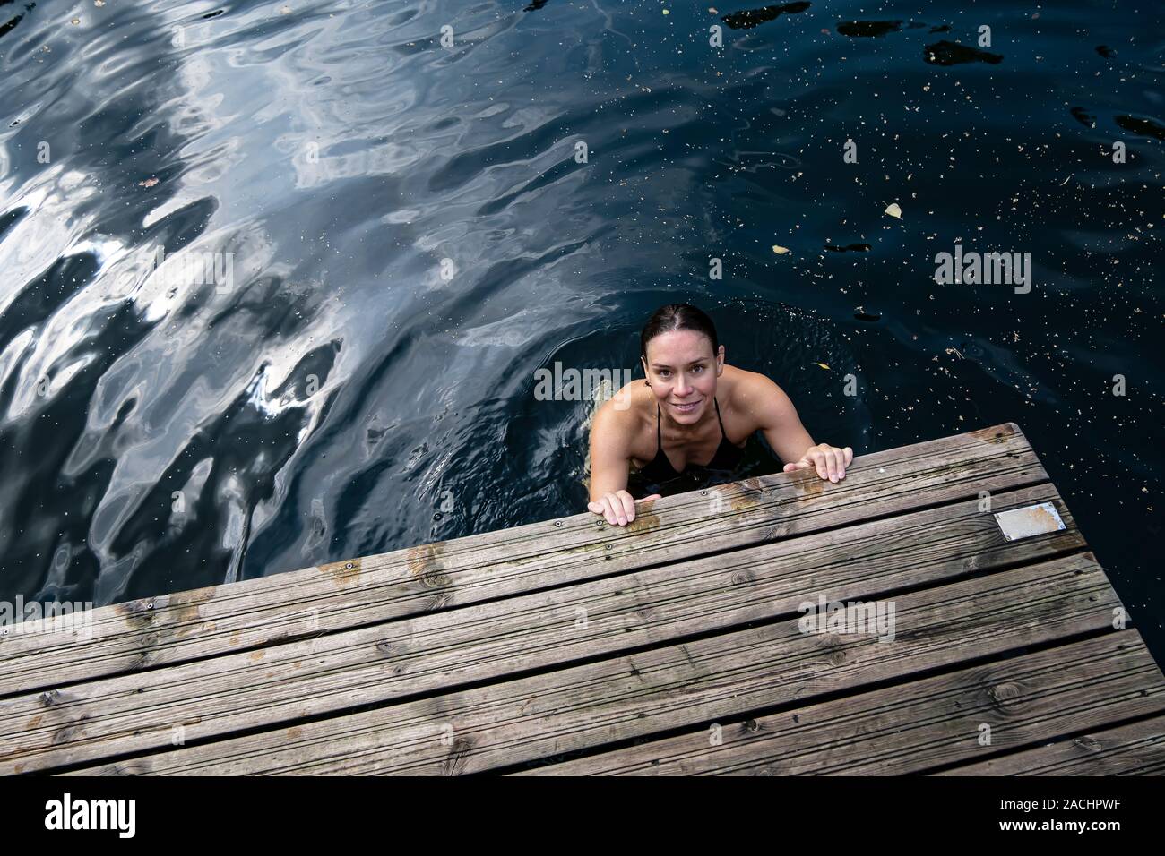 Lächelnde Frau badet in einem klaren See, sie hält ihre Hände auf den Rand eines hölzernen Pier und sieht in die Kamera. Stockfoto