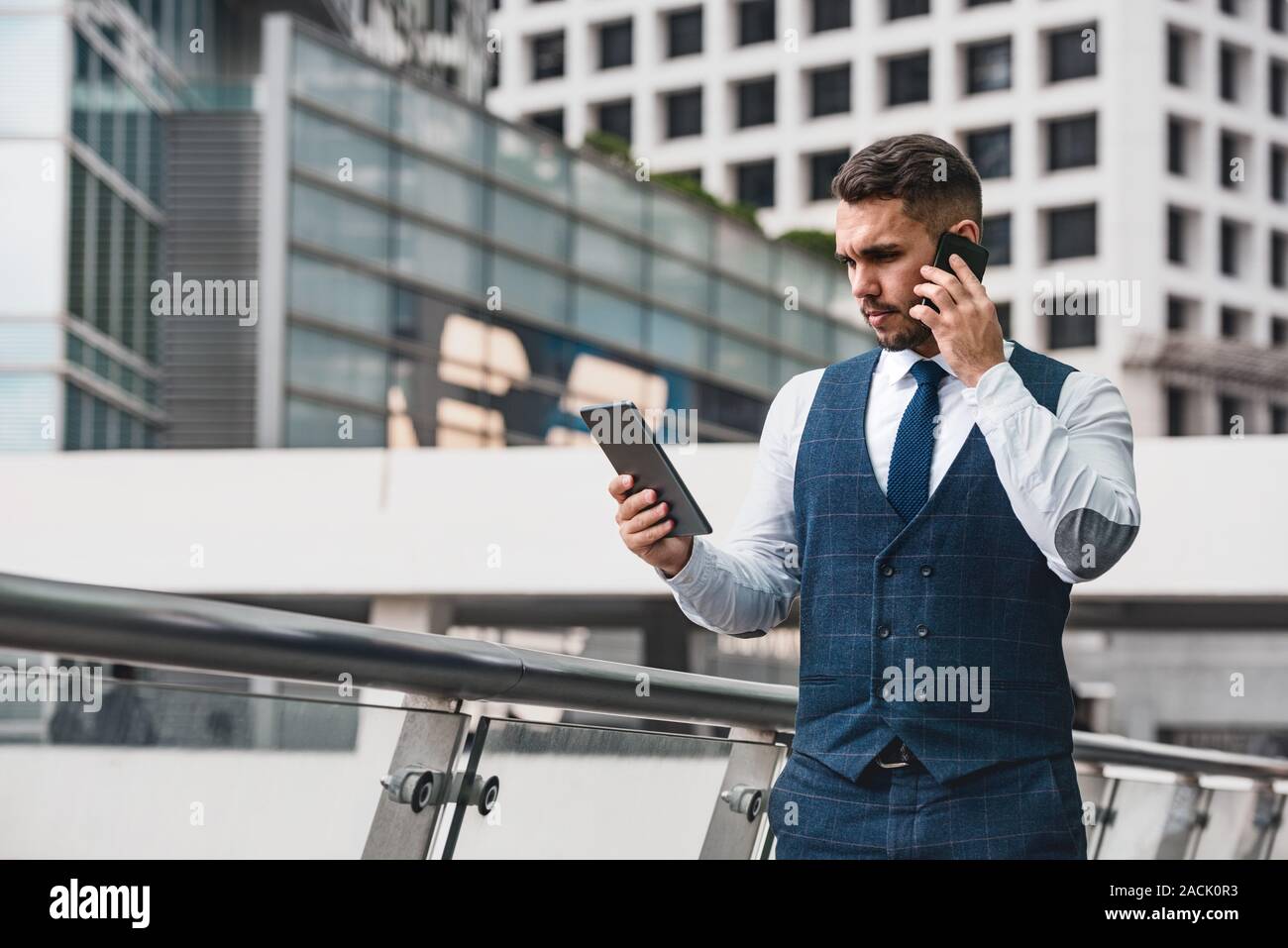 Gut aussehender Geschäftsmann spricht von Mobiltelefon und Blick auf Tablet Computer gegen Big City Stockfoto