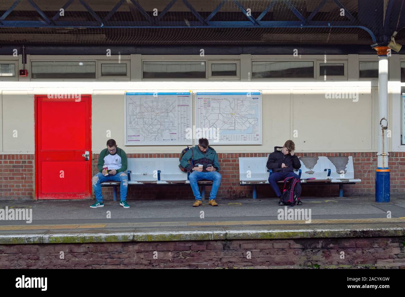 Drei junge Männer sitzen auf einem Sitz mit Blick auf ihre Smartphones beim Warten auf einen Zug bei Kingston Station Surrey, Greater London England Großbritannien Stockfoto