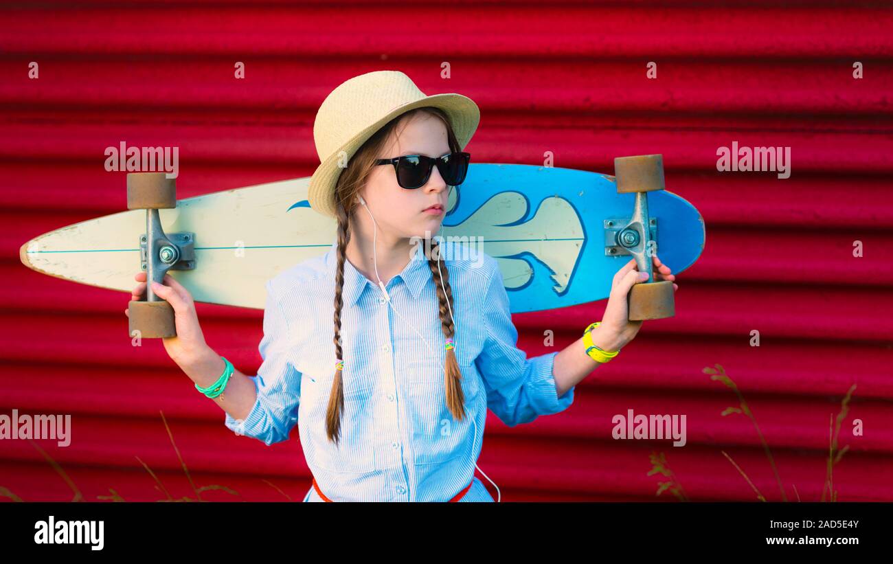 Junge Hipster Mädchen mit Zöpfen in Sonnenbrillen und Strohhut mit einem Longboard auf ihren Schultern - lebendige rote Wand Stockfoto