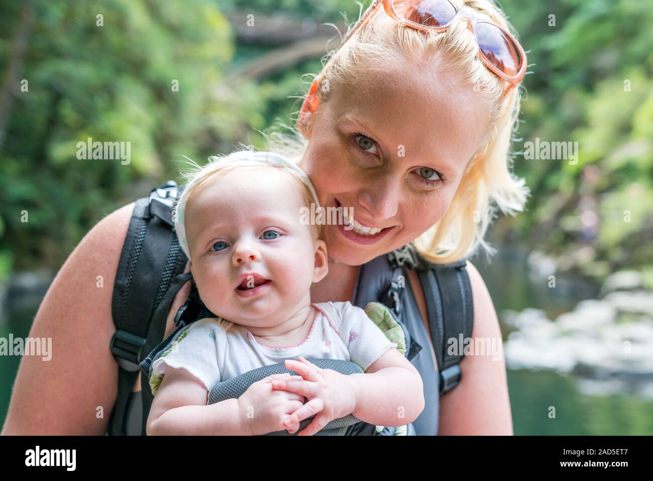 Glücklich lächelnde Mutter mit Baby Kind in der Schlinge, ergonomische Babytrage. Man draußen in der Natur im Sommer. Stockfoto