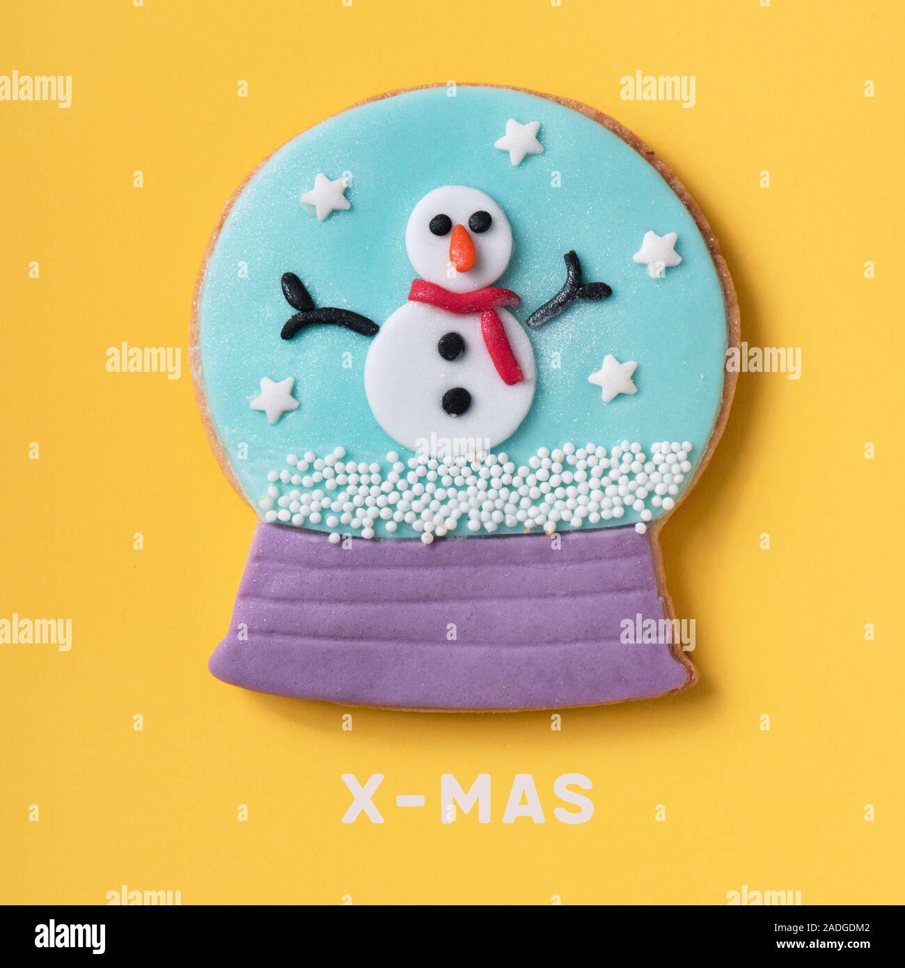 Ein Cookie in Form einer Schneekugel, mit einem Schneemann im Inneren, und der Text x-mas auf gelbem Hintergrund Stockfoto