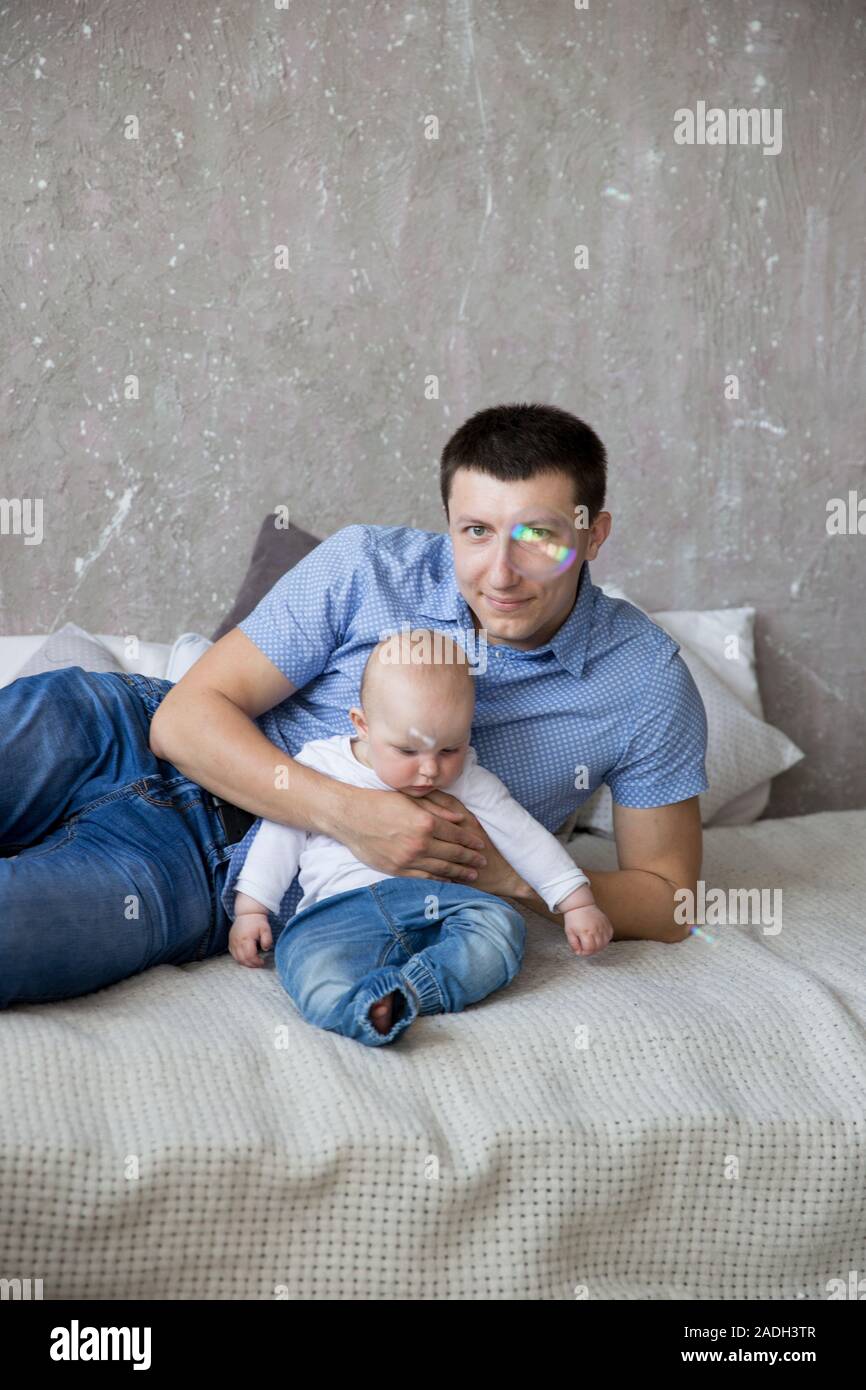 Glückliche Junge kaukasier Familie im Studio. Vater ist das Halten und umarmte seine Tochter in den Händen. Man liegt auf dem Bett. Mann lächelnd. Für Kleinkinder ist ernst. Co Stockfoto