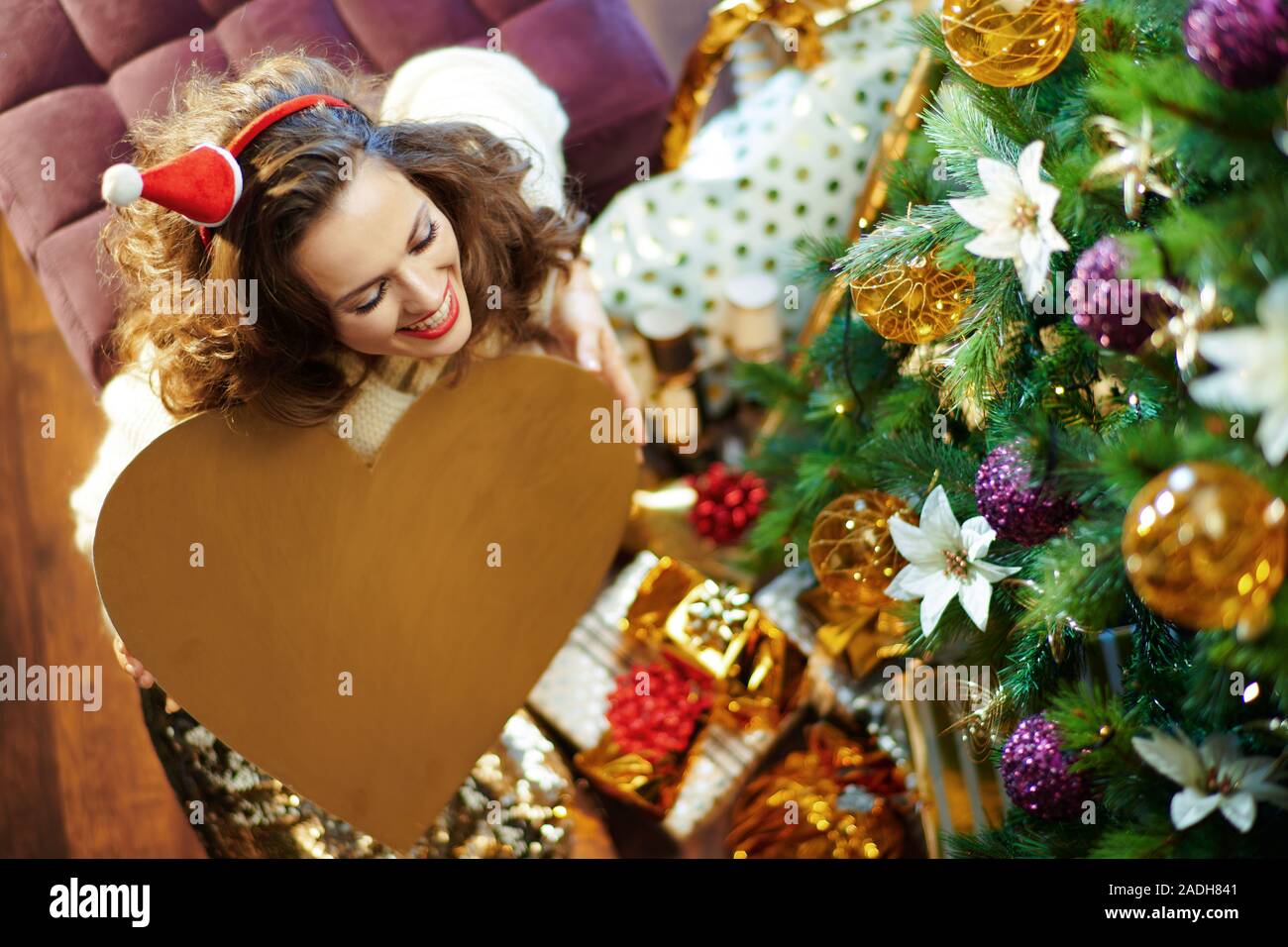 Obere Ansicht von glücklich, elegante Frau mit langen brünetten Haar in Gold Pailletten Rock und weißen Pullover unter dem geschmückten Weihnachtsbaum in der Nähe des heutigen Boxen hol Stockfoto