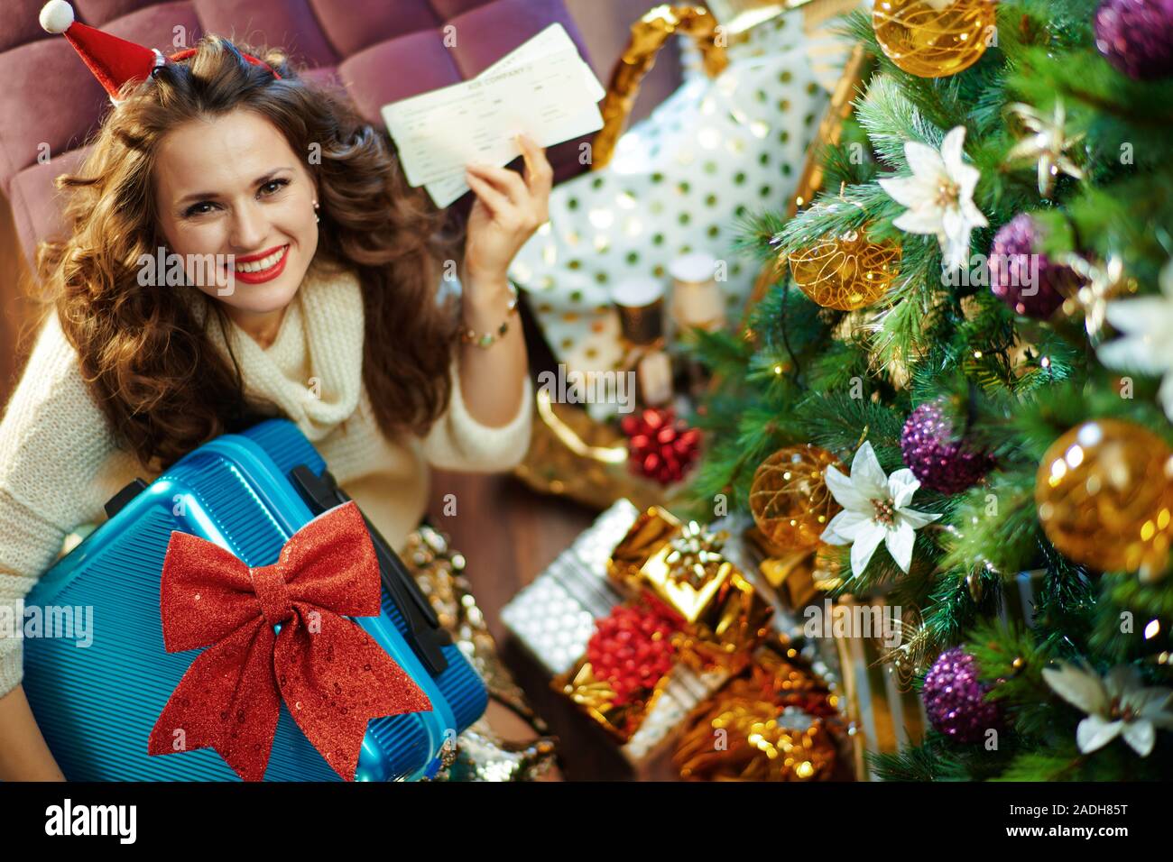 Obere Ansicht der glückliche junge Frau mit langen brünetten Haar in Gold Pailletten Rock und weißen Pullover mit blauen Koffer reisen Flugtickets unter eingerichtet. C Stockfoto