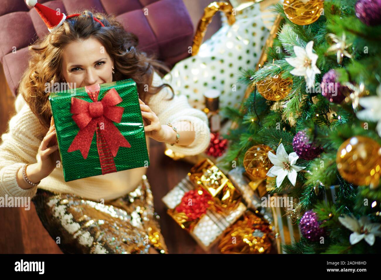 Obere Ansicht von lächelnden jungen Frau mit langen brünetten Haar in Gold Pailletten Rock und weißen Pullover unter dem geschmückten Weihnachtsbaum in der Nähe des heutigen Boxen sho Stockfoto