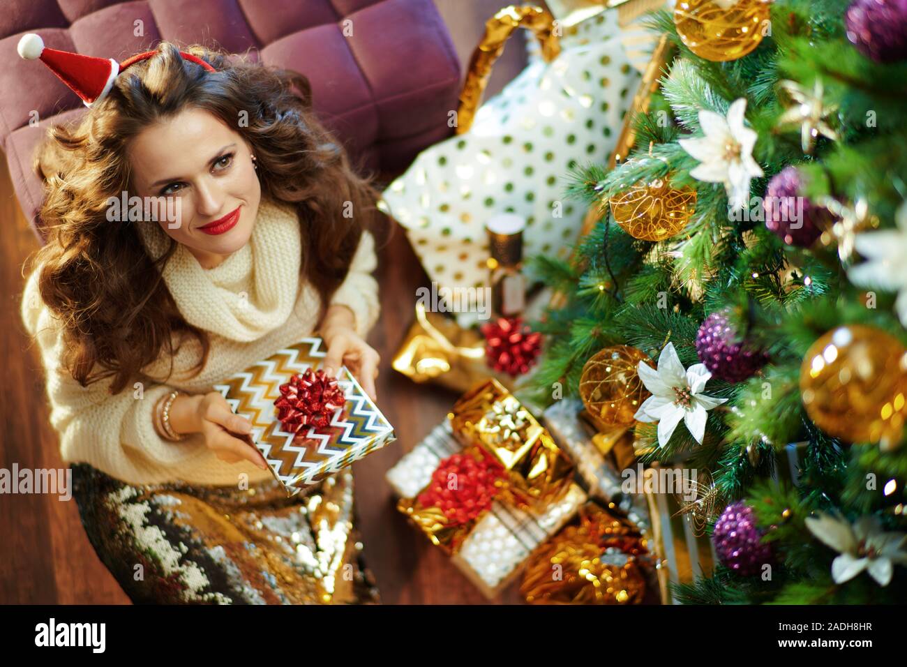 Obere Ansicht, elegante Frau mit langen brünetten Haar in Gold Pailletten Rock und weißen Pullover unter dem geschmückten Weihnachtsbaum in der Nähe des heutigen Boxen Holding w Stockfoto