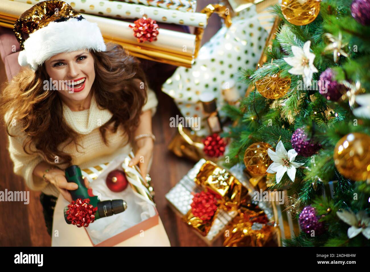 Gerne stilvolle Hausfrau mit langen brünetten Haar in Gold Pailletten Rock und weißen Pullover unter dem geschmückten Weihnachtsbaum in der Nähe des heutigen Boxen wrapping Chri Stockfoto