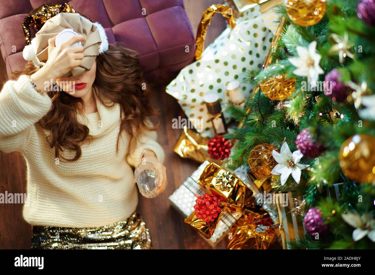 Obere Ansicht der betonten trendy Hausfrau mittleren Alters mit langen brünetten Haar in Gold Pailletten Rock und weißen Pullover mit Glas Wasser Festlegung auf dem Boden Stockfoto