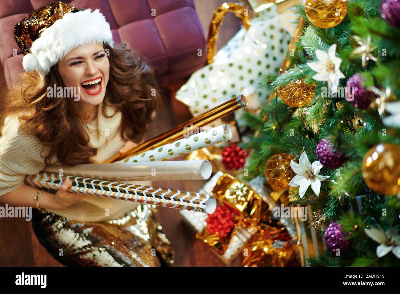 Obere Ansicht von Happy elegante 40-jährige Frau mit langen brünetten Haar in Gold Pailletten Rock und weißen Pullover unter dem geschmückten Weihnachtsbaum in der Nähe von prese Stockfoto