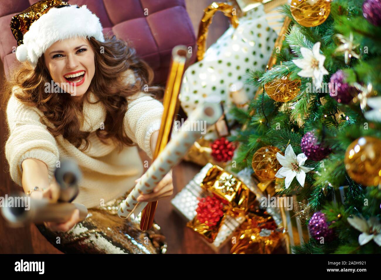 Obere Ansicht von Happy Elegant Hausfrau mit langen brünetten Haar in Gold Pailletten Rock und weißen Pullover unter dem geschmückten Weihnachtsbaum in der Nähe des heutigen Boxen Stockfoto
