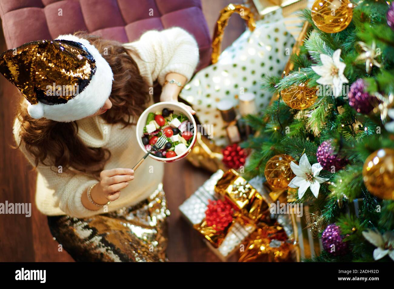 Obere Ansicht der modernen 40-jährige Hausfrau mit langen brünetten Haar in Gold Pailletten Rock und weißen Pullover unter dem geschmückten Weihnachtsbaum in der Nähe des heutigen Stockfoto