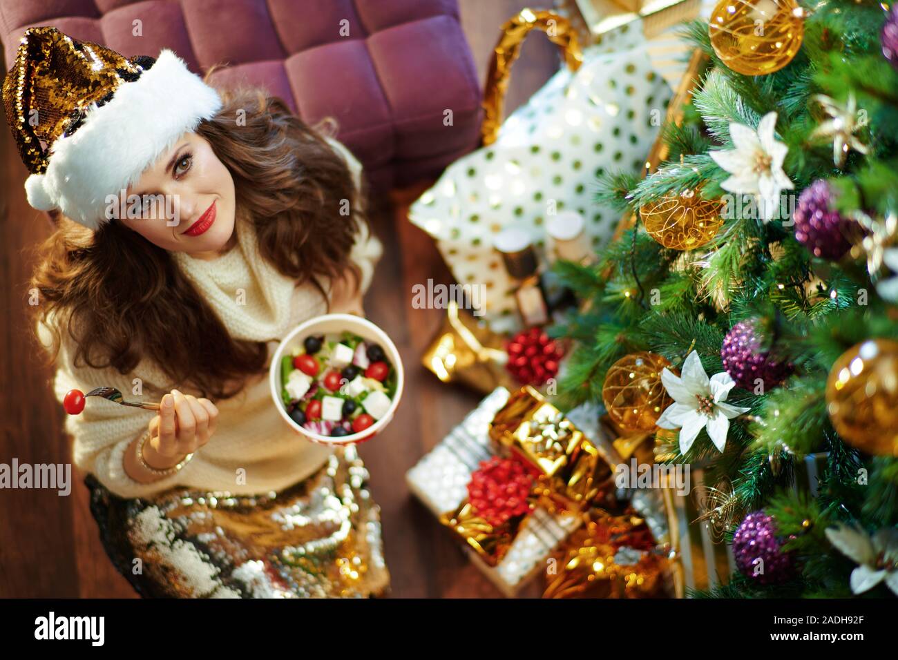 Obere Ansicht von nachdenklich, moderne Frau mit langen brünetten Haar in Gold Pailletten Rock und weißen Pullover unter dem geschmückten Weihnachtsbaum in der Nähe des heutigen Boxen ea Stockfoto