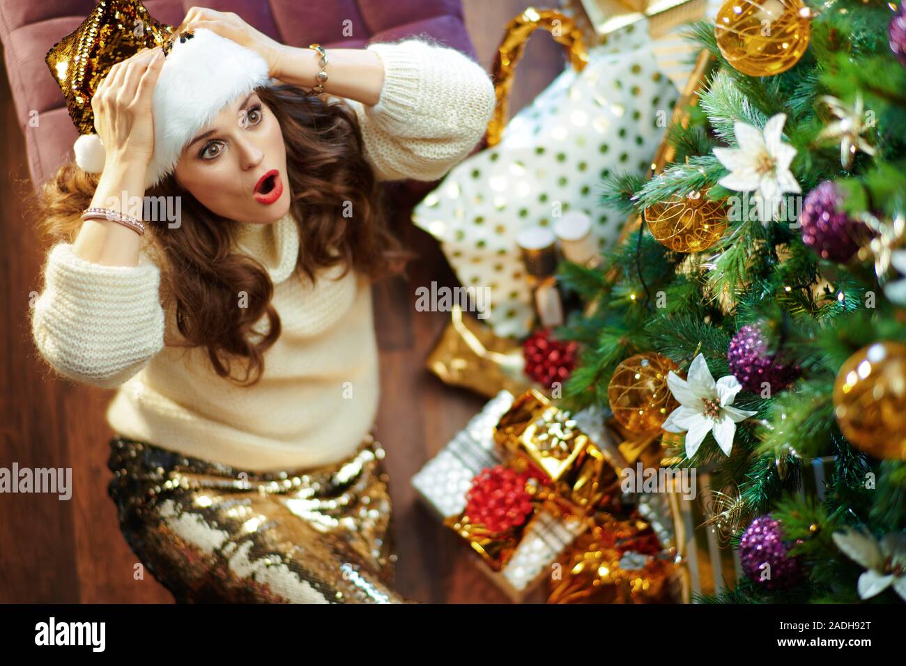 Obere Ansicht von schockiert moderne Frau mit langen brünetten Haar in Gold Pailletten Rock und weißen Pullover unter dem geschmückten Weihnachtsbaum in der Nähe des heutigen Boxen lo Stockfoto