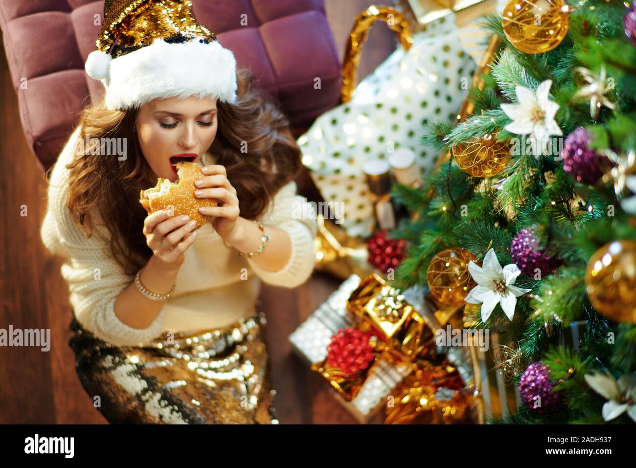 Obere Ansicht von trendigen mittleren Alter Frau mit langen brünetten Haar in Gold Pailletten Rock und weißen Pullover unter dem geschmückten Weihnachtsbaum in der Nähe des heutigen Boxen Stockfoto