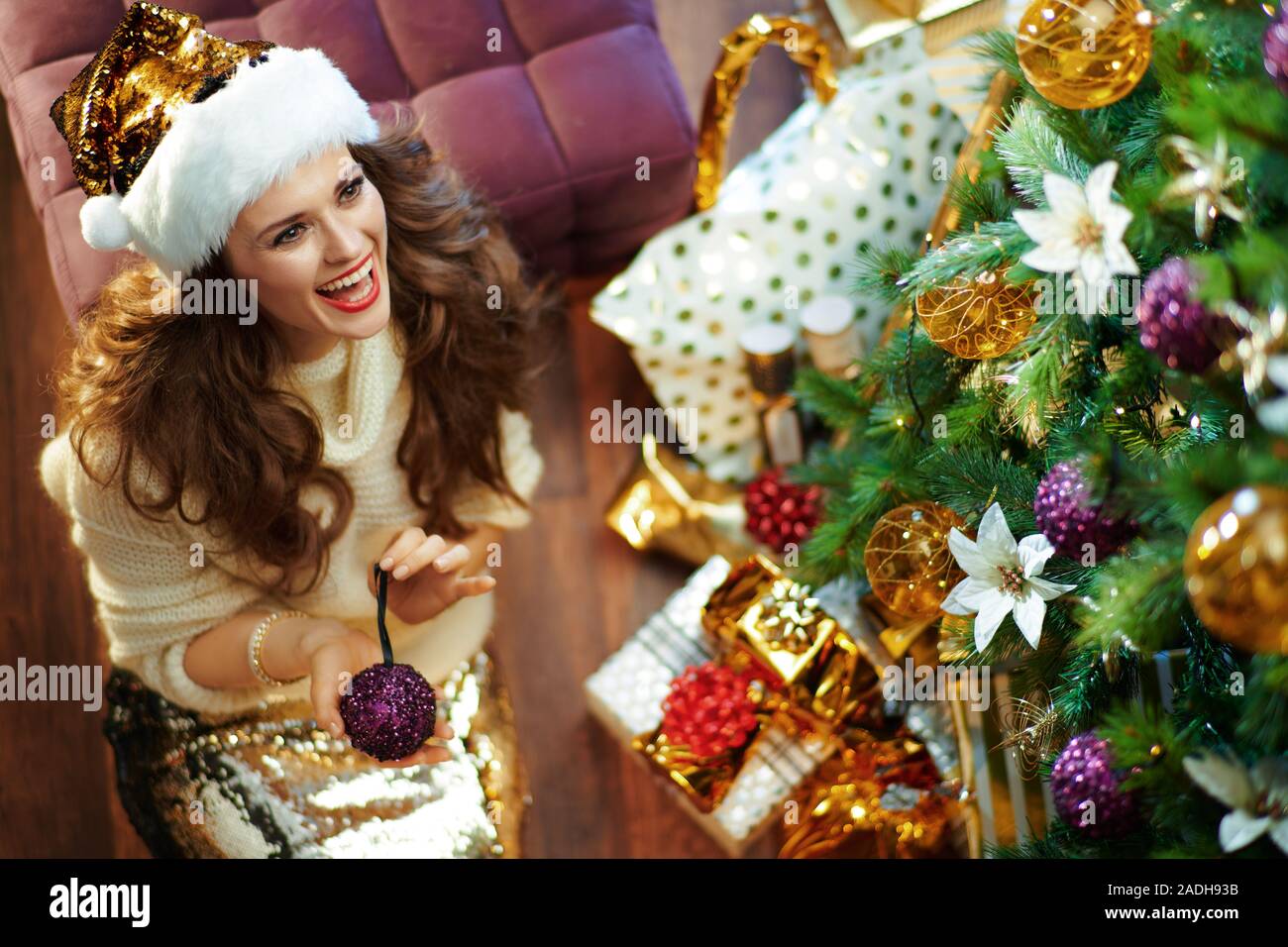 Glücklich, elegante Frau mit langen brünetten Haar in Gold Pailletten Rock und weißen Pullover unter dem geschmückten Weihnachtsbaum in der Nähe des heutigen Boxen suchen Bei copy s Stockfoto