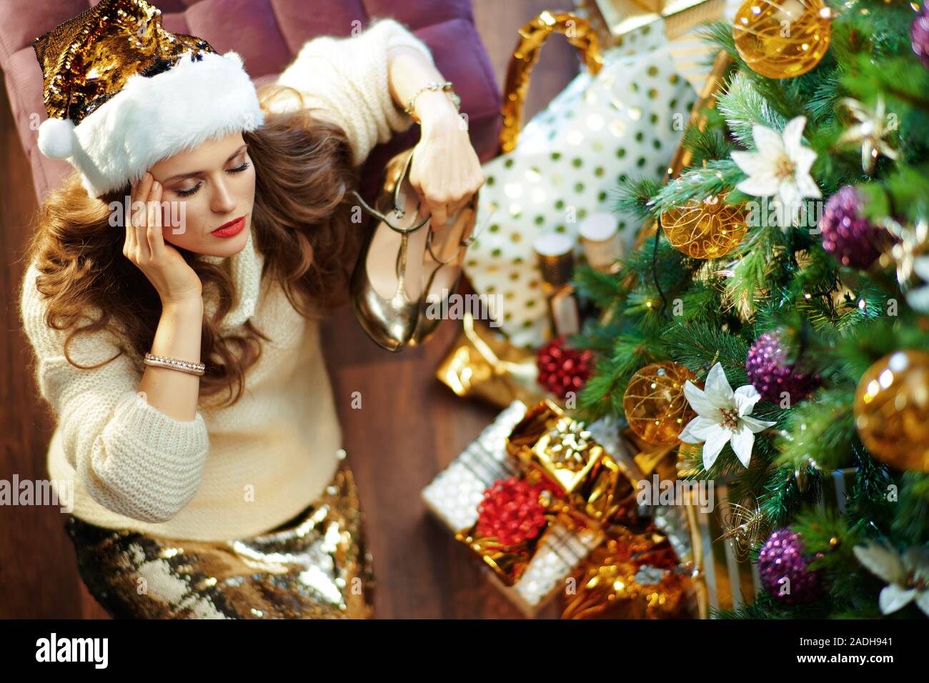 Obere Ansicht der Müde trendy 40 Jahre alte Frau mit langen brünetten Haar in Gold Pailletten Rock und weißen Pullover unter dem geschmückten Weihnachtsbaum in der Nähe von presen Stockfoto