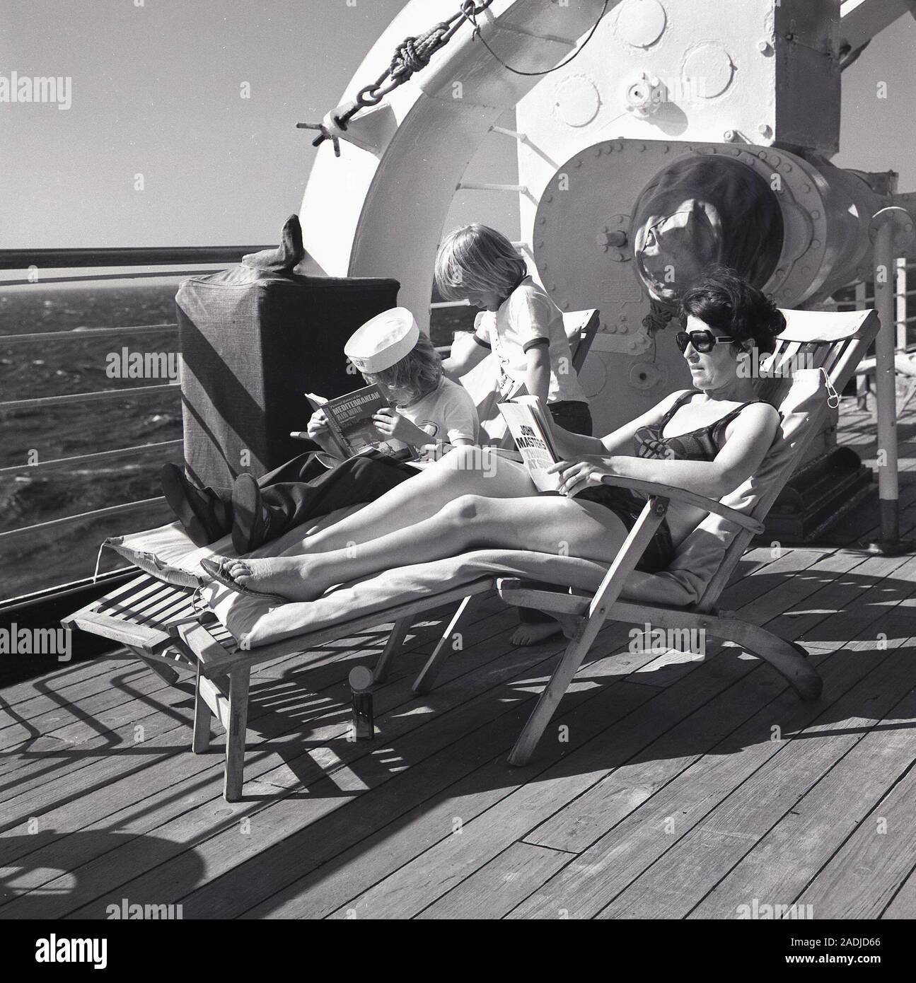 1960, historische, crusiing Stil der 60er Jahre, die in den Ozean, auf dem Deck eines Kreuzfahrtschiffes, eine Frau Passagier auf einem Liegestuhl ein Buch lesen mit ihren beiden Söhnen, die auch lesen. Stockfoto