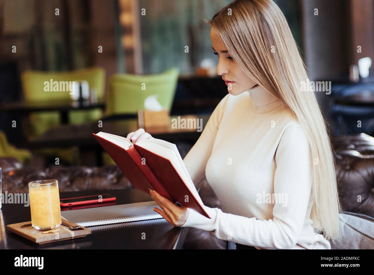 Vorbereitung für die Prüfungen. Blonde Mädchen lesen Sie ein Buch mit rotem Deckel im Restaurant mit Tisch und gelbe Getränk auf es Stockfoto