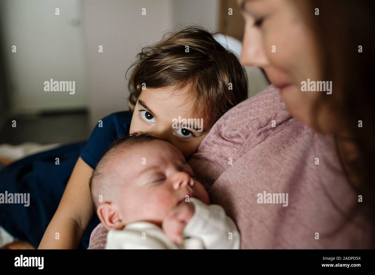 Junges Mädchen mit großen Augen peers hinter Mutter und Neugeborenes Geschwister Stockfoto