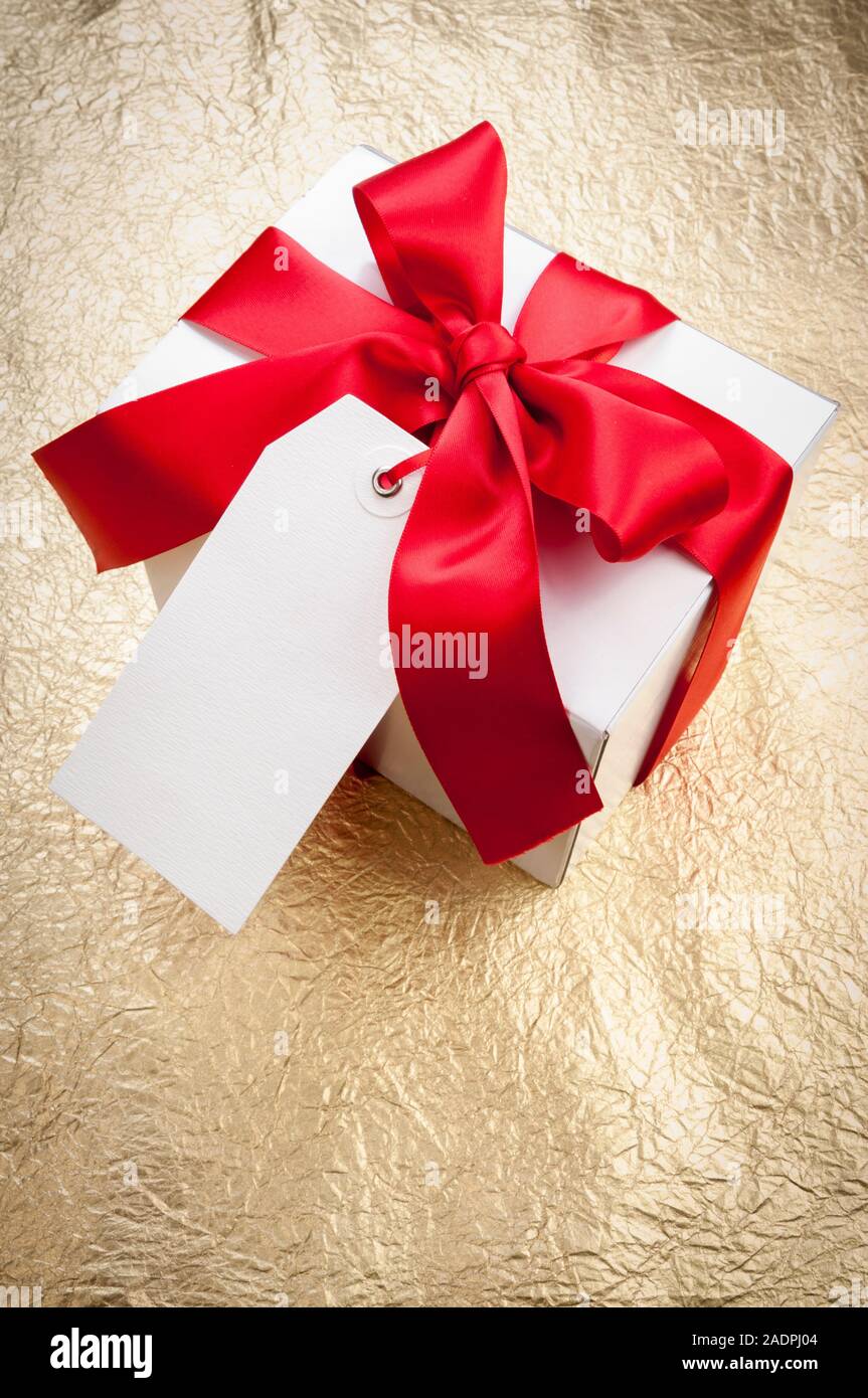Leere weiße Geschenk Tag hängen von einem weißen Kasten mit rotem Satinband auf glänzenden Gold Hintergrund Stockfoto