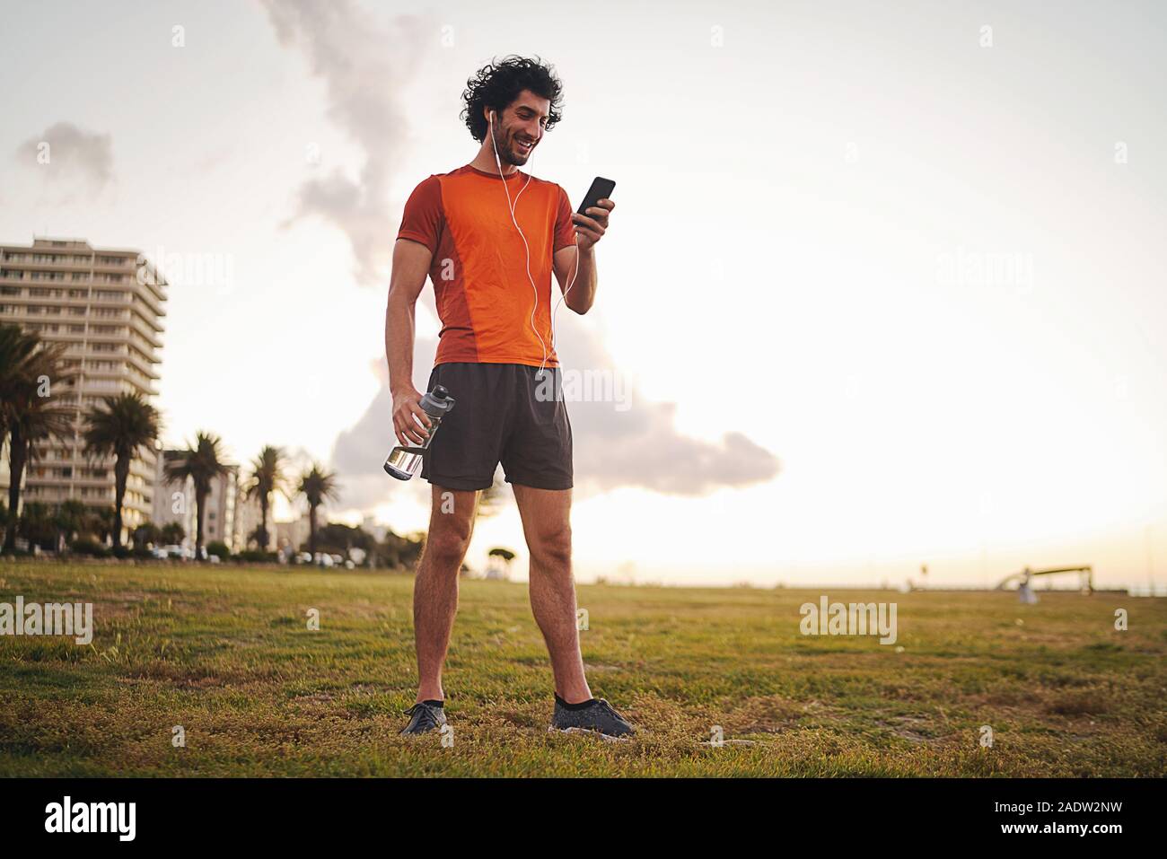 Voller Körper geschossen von einer glücklichen athletischen jungen Mann hält die Flasche in der Hand Musik hören über Kopfhörer mit Smart Phone während im Park stehende Stockfoto