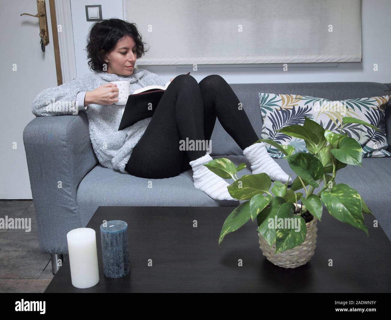 Junge kaukasier Frau mit einem Buch im Schlafanzug, auf dem Sofa sitzen und Kaffee zu trinken. Sie ist zu Hause, gemütliche Konzept. Stockfoto