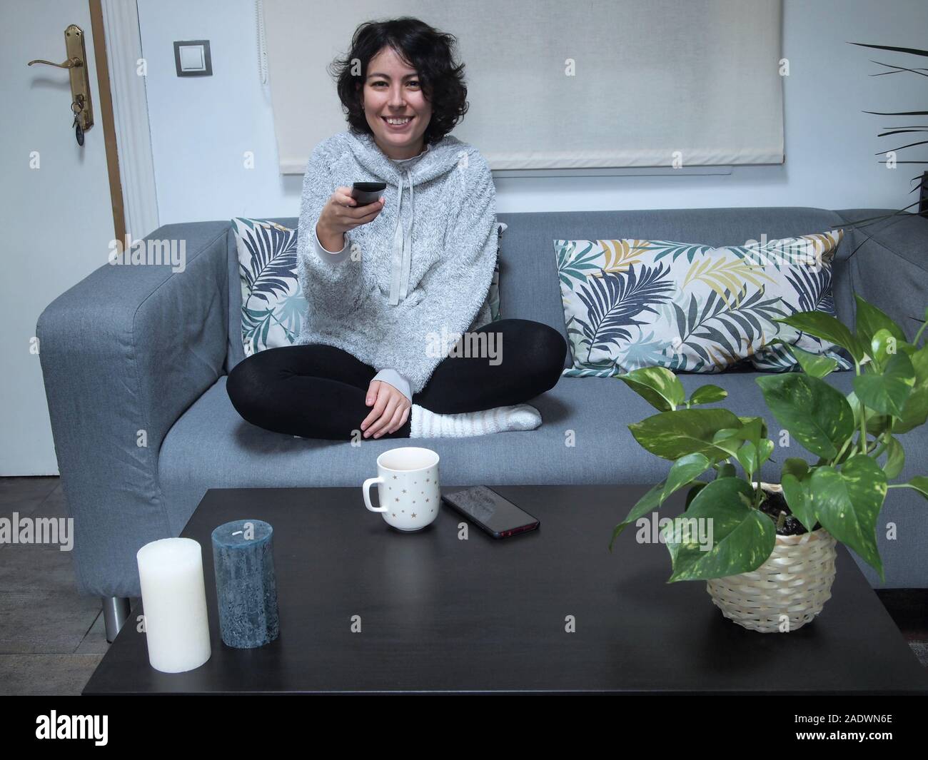 Junge kaukasier Frau lächelnd und mit Hilfe der TV-Fernbedienung im Schlafanzug, sitzen auf dem Sofa mit einer Tasse Kaffee. Sie ist zu Hause, gemütliche c Stockfoto