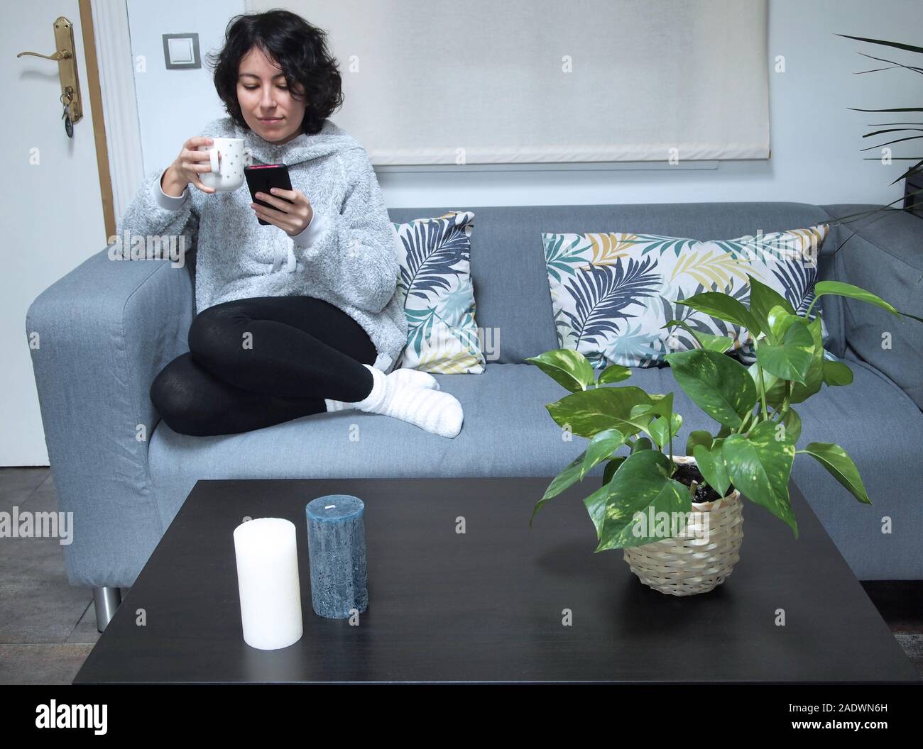 Junge kaukasier Frau auf Ihr Telefon suchen und trinken Kaffee im Schlafanzug auf dem Sofa sitzen. Sie ist zu Hause, gemütliche Konzept. Stockfoto