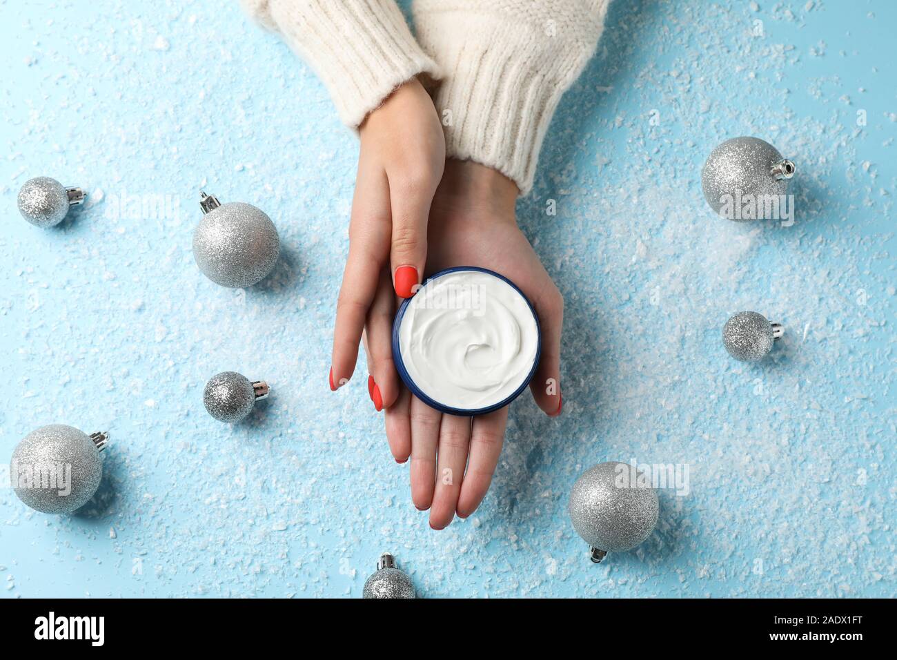 Frau in einem Pullover hält ein Glas Sahne in ihren Händen, feuchtigkeitsspendende Creme für saubere und weiche Haut im Winter, Weihnachten Spielzeug auf verschneiten Blau backgroun Stockfoto