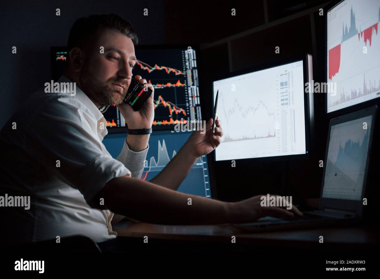 Besetzt Kerl. Bärtiger Mann im weißen Hemd arbeitet im Büro mit mehreren Bildschirmen in Index charts Stockfoto