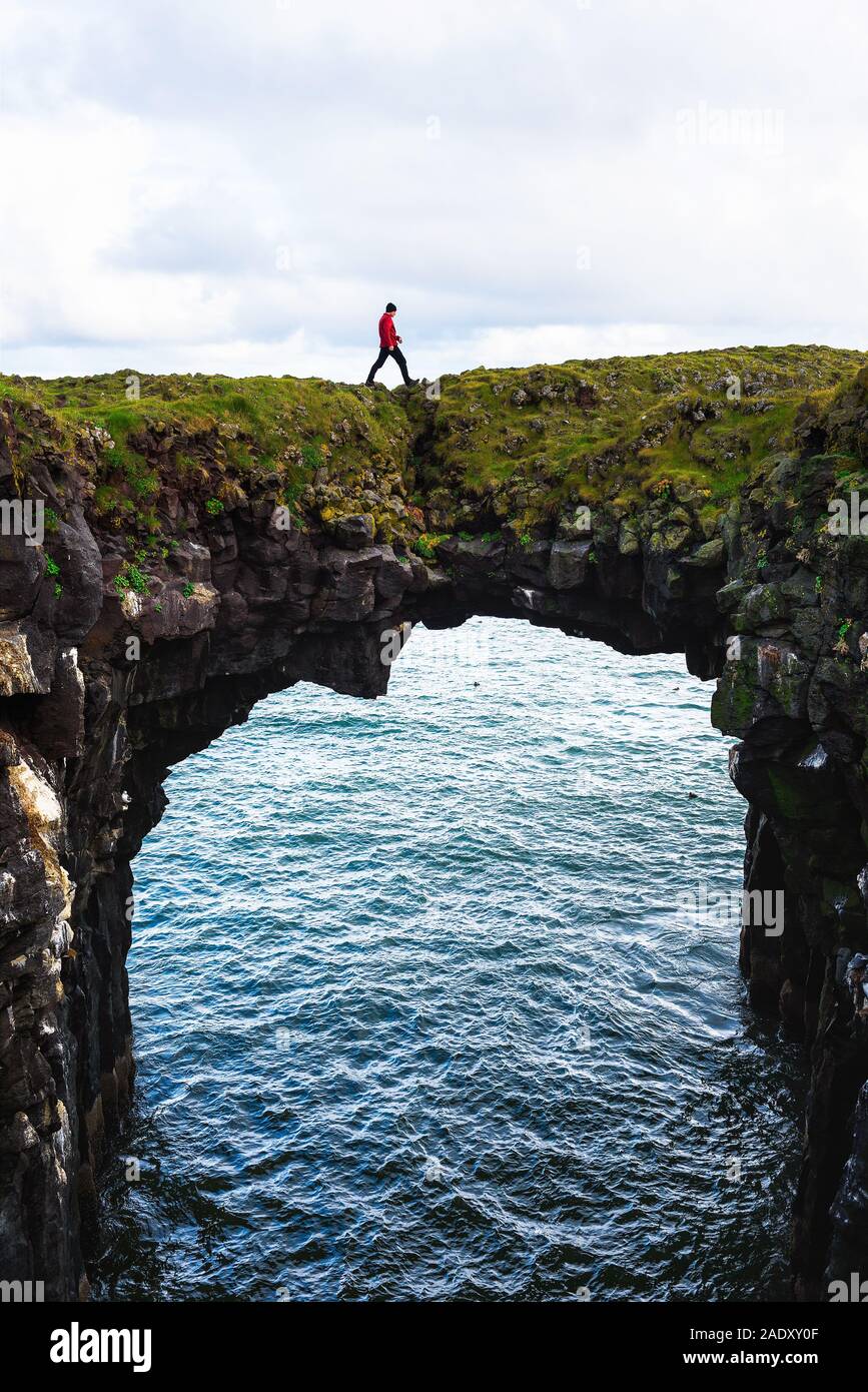 Touristische Spaziergänge über einen natürlichen Felsen Brücke Basaltfelsen in Arnarstapi, Island, mit dem Atlantischen Ozean im Hintergrund. Stockfoto