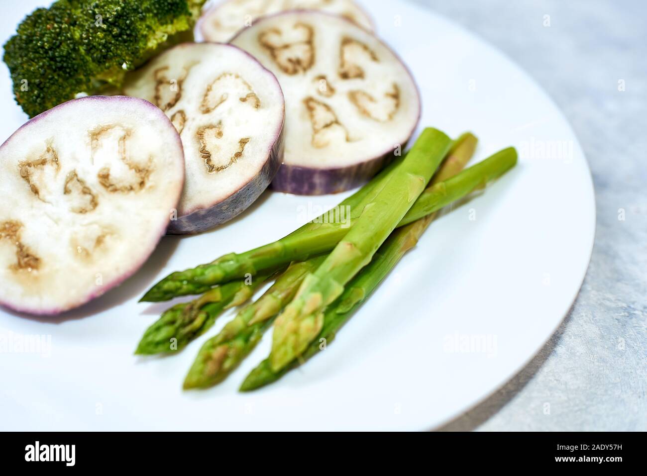 Gedünstetes Gemüse in einer weißen Platte auf einer blauen Tabelle. Auberginen, Brokkoli, Spargel. Stockfoto