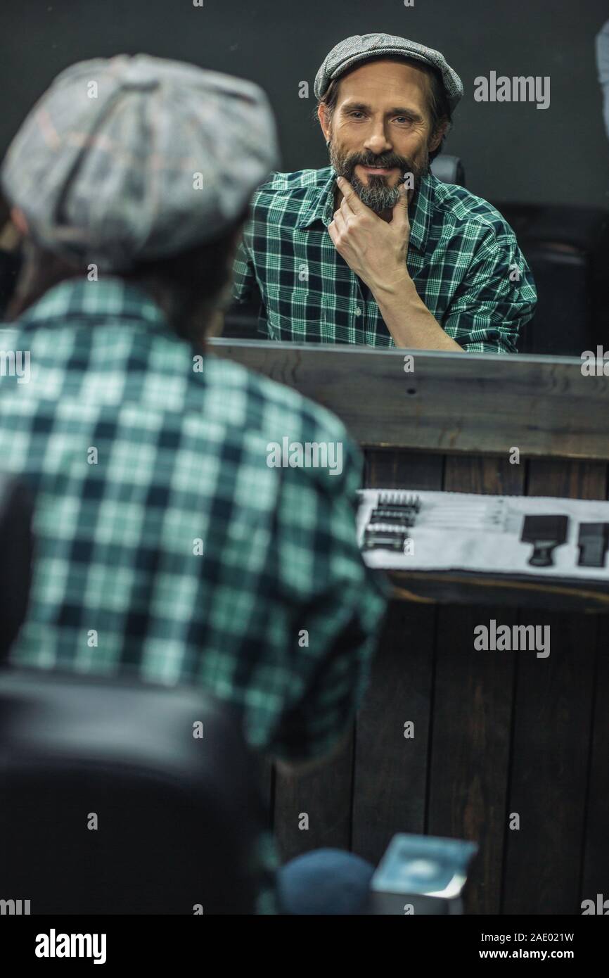 Positiver Mann lächelnd und seinen Bart vor dem Spiegel berühren Stockfoto