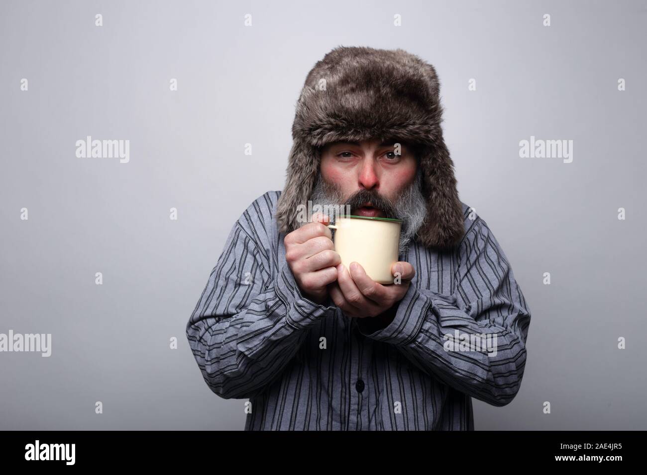 Kranken Mann mit Schlafanzug und mit einer Kappe weht eine heiße Tasse Tee auf einem grauen Hintergrund Stockfoto