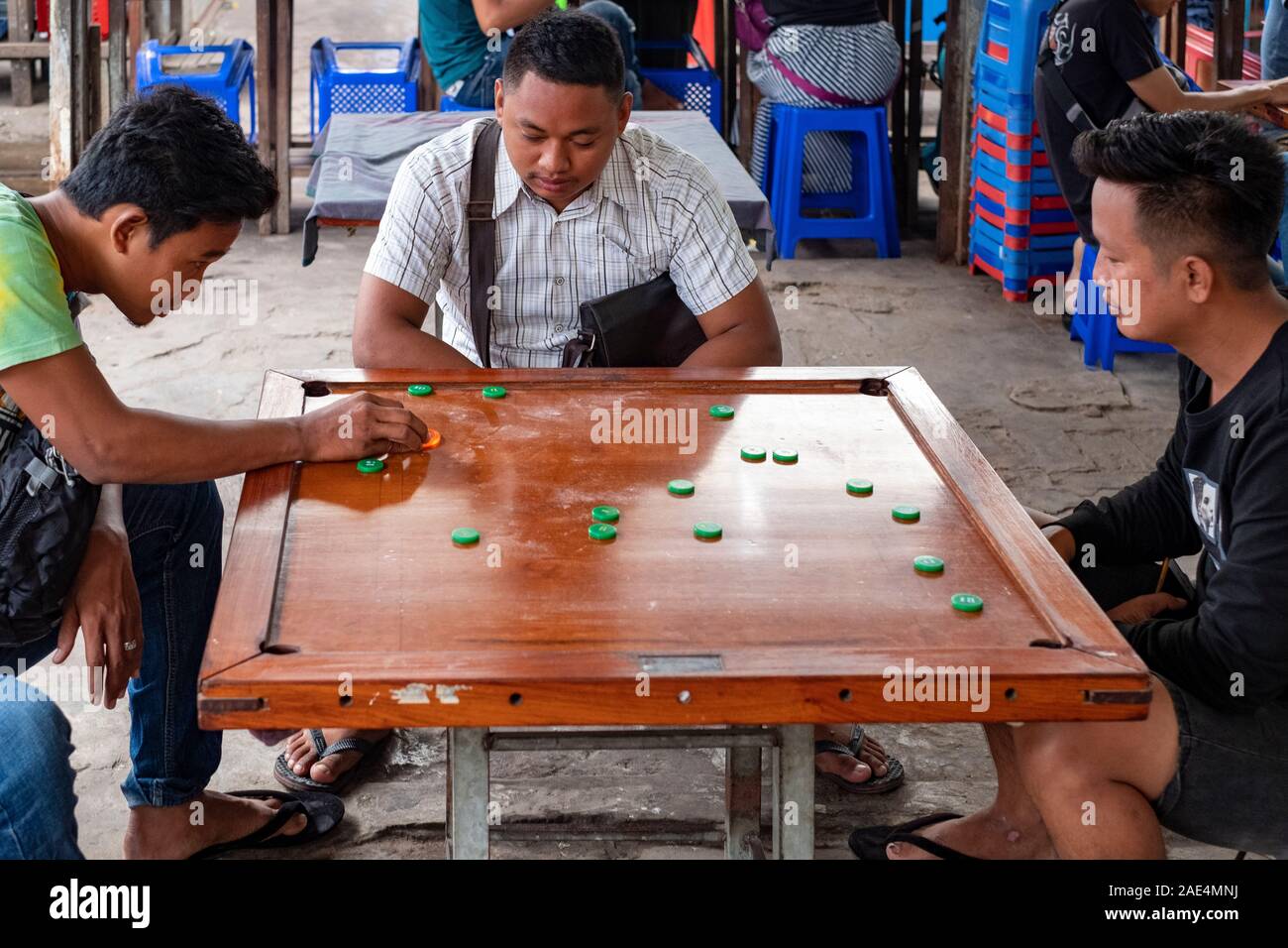 Zwei burmesische Teens spielen das Brettspiel Carrom auf einem Holzbrett mit einem anderen jungen Mann beobachtet das Spiel in einem Markt, in Mandalay, Myanmar (Birma) Stockfoto