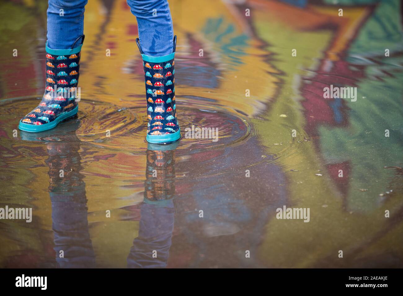 Kind Junge in lustige Gummistiefel in der Pfütze auf der Straße nach Regen. Paar bunte Gummistiefel in einer großen Pfütze mit Graffiti Reflexionen Stockfoto