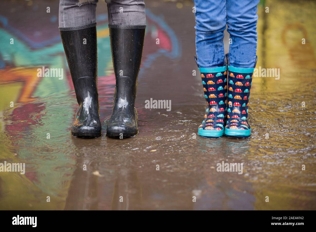 Kind Junge und Frau in lustige Gummistiefel in der Pfütze auf der Straße nach Regen. Familie in bunte Gummistiefel in einer großen Pfütze mit Graffiti Stockfoto
