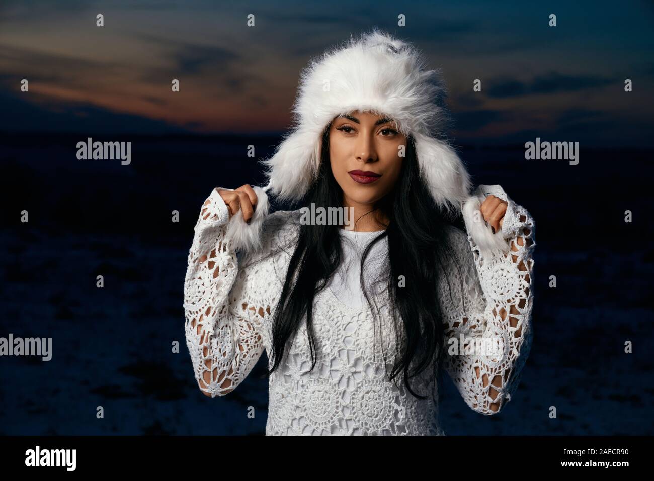 Schöne Mädchen mit schwarzen langen Haaren auf dem Hintergrund der Abendhimmel und Feld mit Schnee. Charmante Modell tragen in Weiß gestrickte Bluse zu berühren weiß warme Mütze. Stockfoto
