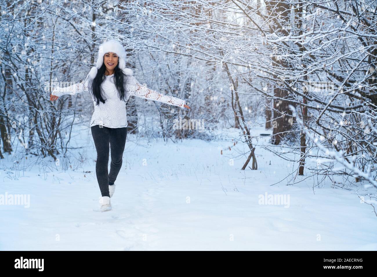 Schöne Mädchen in der modernen Kleidung wandern im Winter Wald. Positive, verspielte Dame in warmen weißen Hut, aus Gewirken Bluse, Jeans und Turnschuhe auf dem Hintergrund der Bäume mit Schnee posieren. Stockfoto