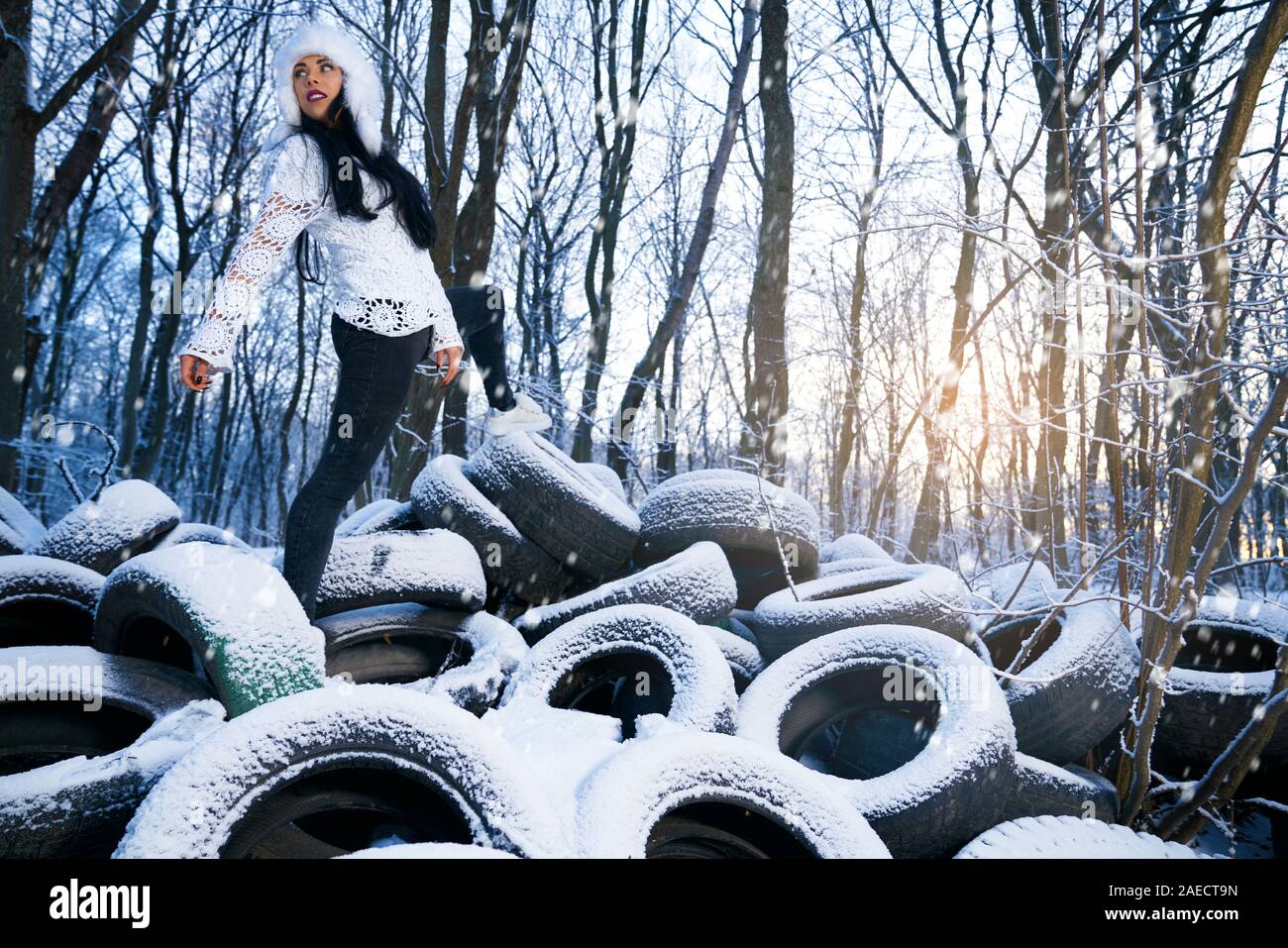 Stilvolle Modell auf alten Reifen Dumping im Wald posieren. Schöne Mädchen tragen in Weiß warme Mütze und weiße Bluse, Jeans und Turnschuhe auf PKW-Reifen auf Schnee. Stockfoto